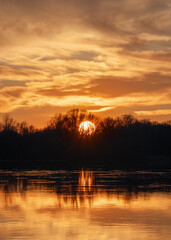 Scenic orange sunset with clouds over forest and river. The Sun hides behind the trees