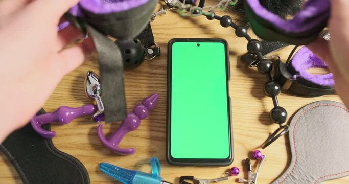 Men's hands place sex handcuffs on table, smartphone with green screen surrounded by sex toys. Concept of online adult app, anonymous sex shop.