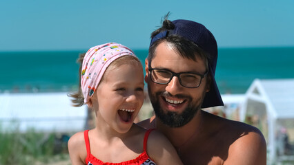 Fototapeta premium Happy father and daughter look at camera and laugh on background of hotel beach with blue sea. Concept of summer vacation, travel, holiday. People outdoors on seascape