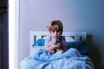 Little calm boy sit in bed hold plush toy covered with blanket