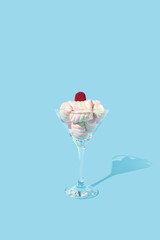 Raspberry gummy candy on the top of pile of marshmellows in cocktail glass on pastel blue background. Sweet food and drink concept.