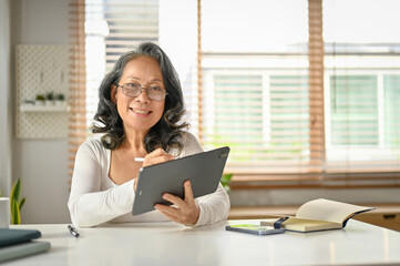 Smiling and happy 60s Asian-aged woman using her digital tablet at her home workspace