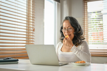 Chilling 60s Asian-aged woman eating cookies while using her laptop at her home workspace.