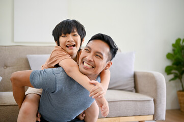 Joyful and happy Asian dad playing with his little son in the living room, piggyback