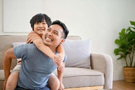 Joyful and happy Asian dad playing with his little son in the living room, piggyback