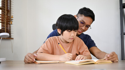 Naklejka premium Cute Asian little boy focusing on his homework while his dad is teaching and helping.