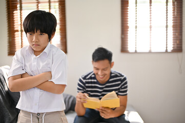 An angry Asian boy stands with his arms crossed, feeling lonely by his father