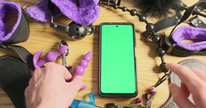Smartphone with green screen lies on table among the sex toys. Man's finger clicks on screen holding anal stimulator.