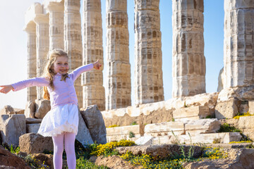 A happy, blonde tourist girl in a white summer dress in front of the Temple of Poseidon in Sounion,...