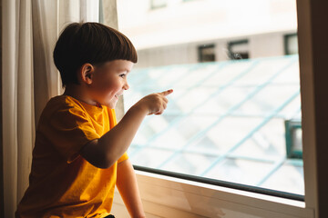 Little cute attractive boy sitting on windowsill and look at window, boy point his finger on...