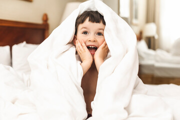 Little boy wrapped himself in a white blanket while sitting on the bed and smiling. Happy kid look surprised, his hands on cheeks, amazement toddler boy. Good morning.