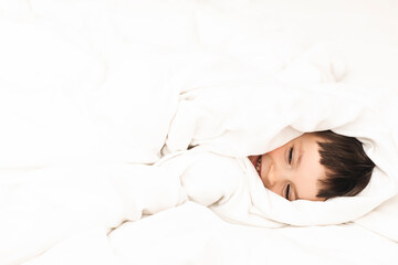 Little cute boy wrapped himself in a white blanket while lying on the bed and smiling. Happy kid look happy, amazement toddler boy. Good morning.