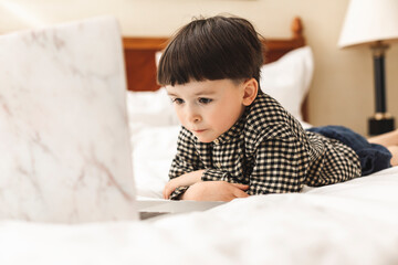 Fototapeta premium Smart looking preschool boy lying on the bed using notebook computer in his room looking at the screen. Online learning, Home preschool, New normal, Home Based Learning