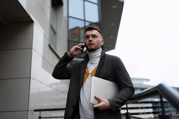 successful european young businessman talking on the phone holding a laptop in his hands on the background of the business center