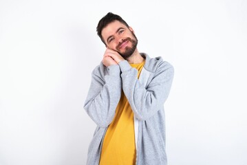 Charming serious Young caucasian mán wearing trendy clothes over white background keeps hands near face smiles tenderly at camera