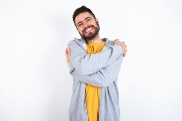 Charming pleased Young caucasian mán wearing trendy clothes over white background embraces own body, pleasantly feels comfortable poses. Tenderness and self esteem concept