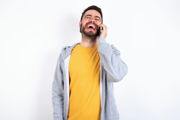 Funny Young caucasian mán wearing trendy clothes over white background laughs happily, has phone conversation, being amused by friend, closes eyes.