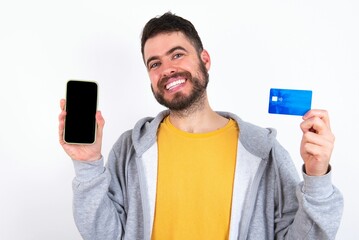 Young caucasian mán wearing trendy clothes over white background opened bank account, holding smartphone and credit card, smiling, recommend use online shopping application