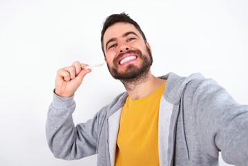 Young caucasian mán wearing trendy clothes over white background make selfie holding an invisible braces aligner, recommending this new treatment. Dental healthcare concept.