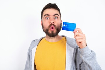 Photo of Young caucasian mán wearing trendy clothes over white background amazed shocked hold credit card payment