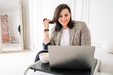 A female psychologist consultant online uses a laptop in business clothes confident work in the office sitting in an armchair. A happy person at work every day.