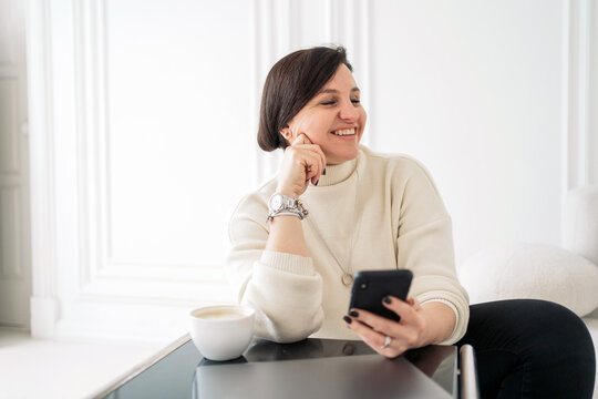 A Woman In Business Clothes Has A Confident Job In The Office Using An Online Phone. A Satisfied Person Is In The Office Every Day, Drinking Coffee.