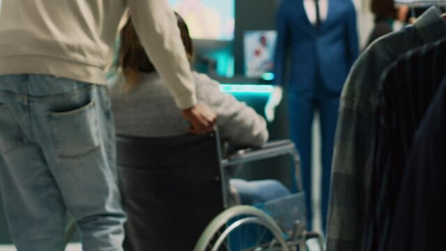 Female client with disability coming to buy clothes, looking to increase wardrobe in clothing store. Young woman wheelchair user shopping for modern trendy wear, market boutique. Handheld shot.