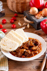 Pork goulash meat with dumplings on plate, cutlery, garlic, onion, pepper, tablecloth in the background - typical Czech food