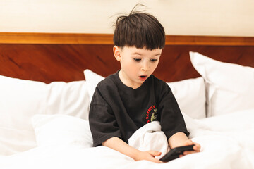 Caucasian boy in t-shirt is looking surprised by something on the phone. His mouth is wide open and so his eyes. Boy sitting on the bed in bedroom. Kid scared in shock with a surprise face, afraid.