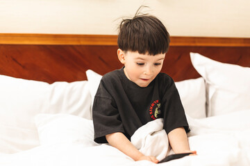 Caucasian boy in t-shirt is looking surprised by something on the phone. His mouth is wide open and so his eyes. Boy sitting on the bed in bedroom. Kid scared in shock with a surprise face, wow.