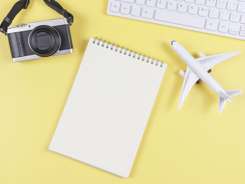 flat lay of airplane model, computer keyboard, opened blank page notebook and digital camera with copy space on yellow background, business and traveling concept.