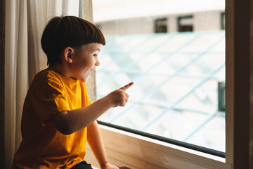 Little cute attractive boy sitting on windowsill and look at window, boy point his finger on something, what he saw on window. Kid look shocked amazement and surprised, he wear yellow t-shirt at home.