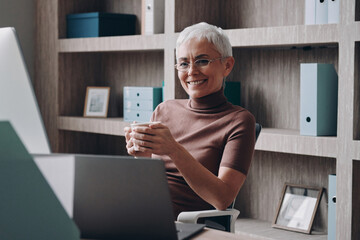 Happy senior businesswoman enjoying hot drink while looking at computer monitor in the office