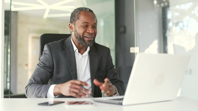 Smiling Mature African American Man In Formal Suit Talking On Video Call Using Laptop While Sitting At Workplace In Modern Office. Happy Middle Aged Businessman With Emotional Having An Online Chat