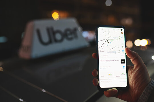 Uber app displayed on smartphone held in hand in front of Uber taxi sign on top of a car at night with city lights background with soft focus. Taking a cab concept. Warsaw, Poland - October 23, 2021