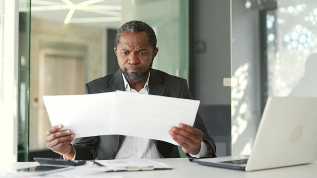 Confused mature businessman having difficulty with paper work sitting at workplace in modern office. The accountant looks at the documents, spreads his hands and cannot understand the problem
