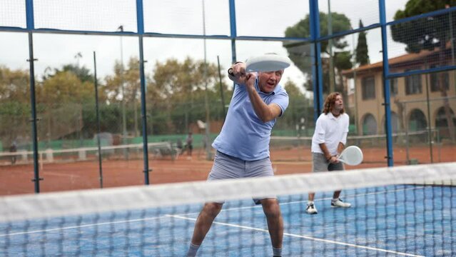 Portrait of active emotional elderly man playing padel tennis on open court on warm autumn day, swinging racket to return ball over net 