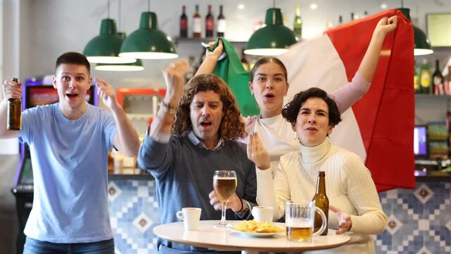 Emotional young adult sports fans waving Italian flag while drinking beer and watching game match together in bar
