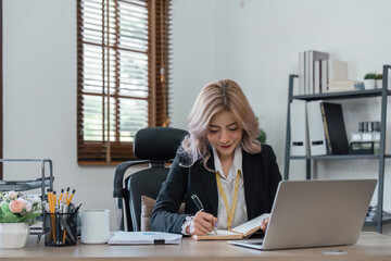 Portrait of smiling business woman in sitting at desk, using laptop and writing in notebook tutorial