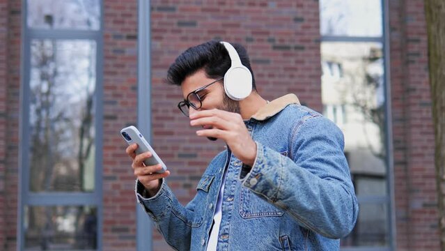 Happy cool indian man wearing headphones and glasses dancing alone on street. Smiling young positive mixed race guy hipster listening music standing in city outdoors, feeling free and funky.