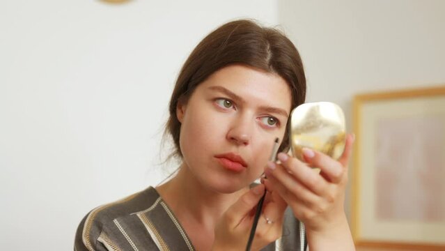 Young woman looks in the mirror, holds a brush and applies shadows on the eyelids. Prepare getting ready in the morning, smiling beautiful girl doing make up put cosmetics on face
