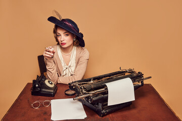 Attractive woman writer wearing old-fashioned clothes sitting at table and smoking over beige background - obrazy, fototapety, plakaty