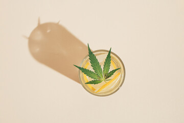 Sunlit glass of water with ice, lemon slices and marijuana leaf against bright sunny background. Minimal summer concept. Top view.