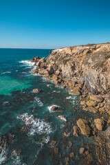 Fototapeta na wymiar Breathtaking cliffs with crashing waves in the afternoon sun on the Atlantic coast near Vila Nova de Milfontes, Odemira, Portugal. In the footsteps of Rota Vicentina. Fisherman trail. Clear blue sky