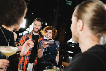 Smiling people holding cocktails near blurred interracial friends in bar at night.