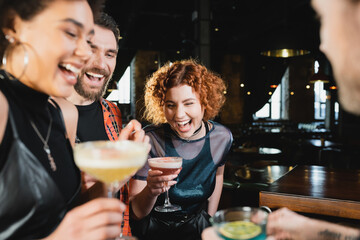 Carefree redhead woman spending time with interracial friends holding cocktails in bar.
