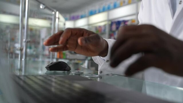 This 4K high close-up quality stock footage showcases a hands of knowledgeable dark-skinned male pharmacist in a white coat working on a laptop in slo mo. Healthcare and medicine in a retail drugstore