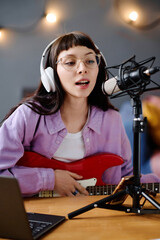 Vertical image of singer with guitar recording her song, singing in microphone at table