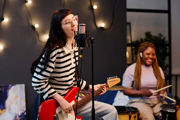 Young woman playing guitar and singing while rehearsing with her friend playing drums in background
