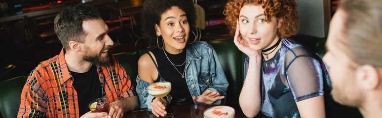 Excited interracial women holding cocktails near blurred men in bar, banner.